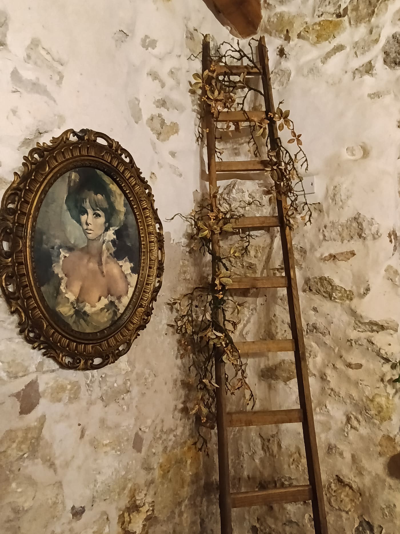 Quadro ovale in cornice barocca dorata accanto a una scala a pioli antica, su muro in pietra viva