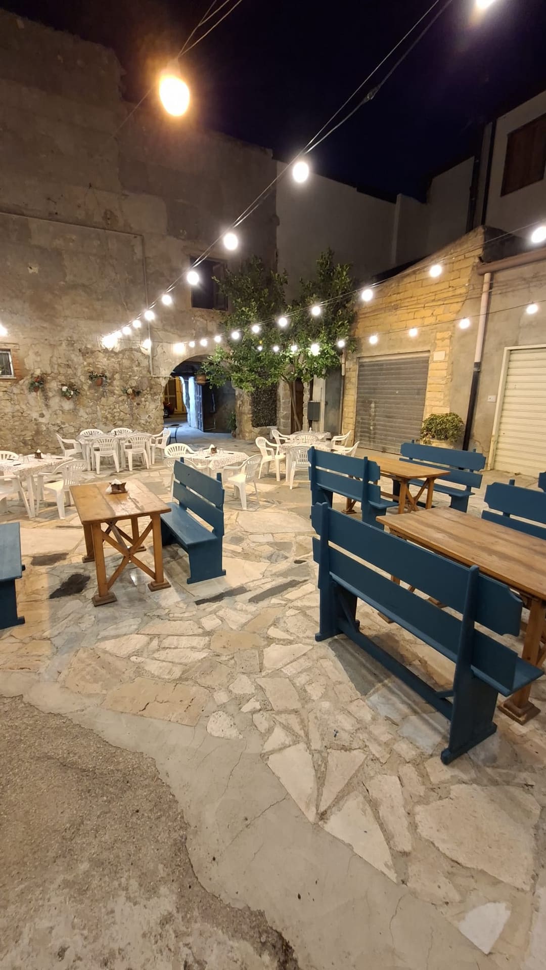 Il cortile di notte con tavoli in legno, panche blu e tavoli apparecchiati sotto i festoni di lucine, sullo sfondo un albero di agrumi