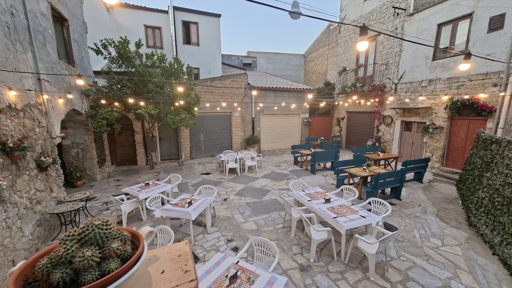 Il cortile in pietra di Sicilia in Bocca al tramonto: tavoli apparecchiati con tovaglie a quadri, panche blu, bouganville in fiore sui balconi e festoni di lucine accese