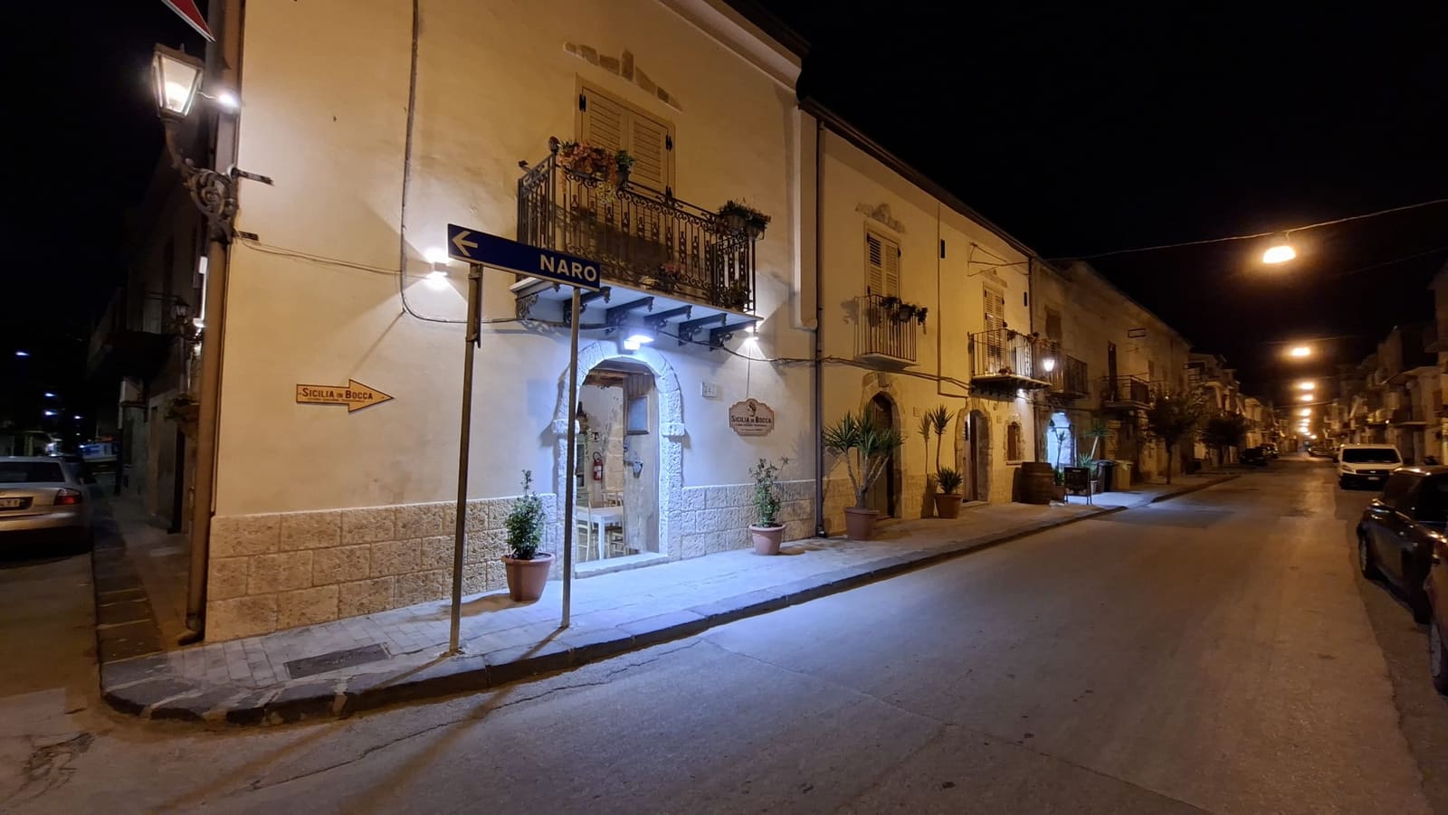 La facciata di Sicilia in Bocca di sera sull'angolo di Corso Garibaldi: palazzo color crema con balconi in ferro battuto, portale ad arco illuminato, insegna in legno e una freccia che indica l'ingresso