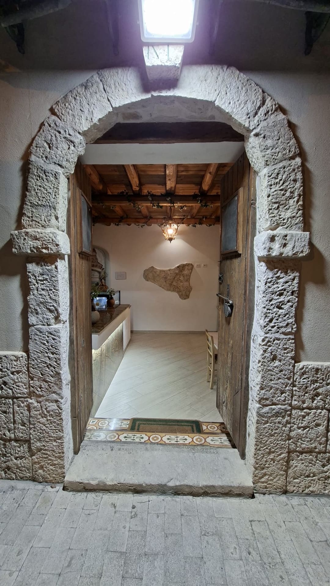 Il portale ad arco in pietra aperto sulla sala: soffitto a travi di legno, lanterna in ferro battuto, bancone e la lastra di pietra a forma di Sicilia appesa alla parete