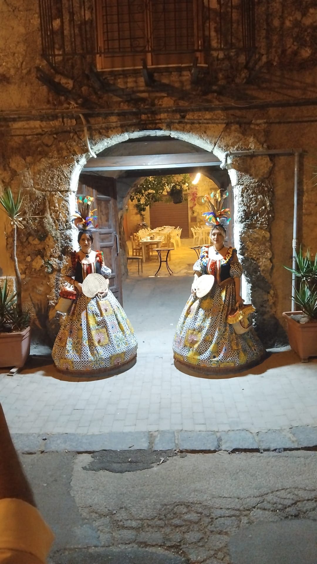 L'ingresso ad arco in pietra di Sicilia in Bocca di sera, con due figuranti in abiti tradizionali siciliani colorati e piumati ad accogliere gli ospiti; sullo sfondo il cortile illuminato
