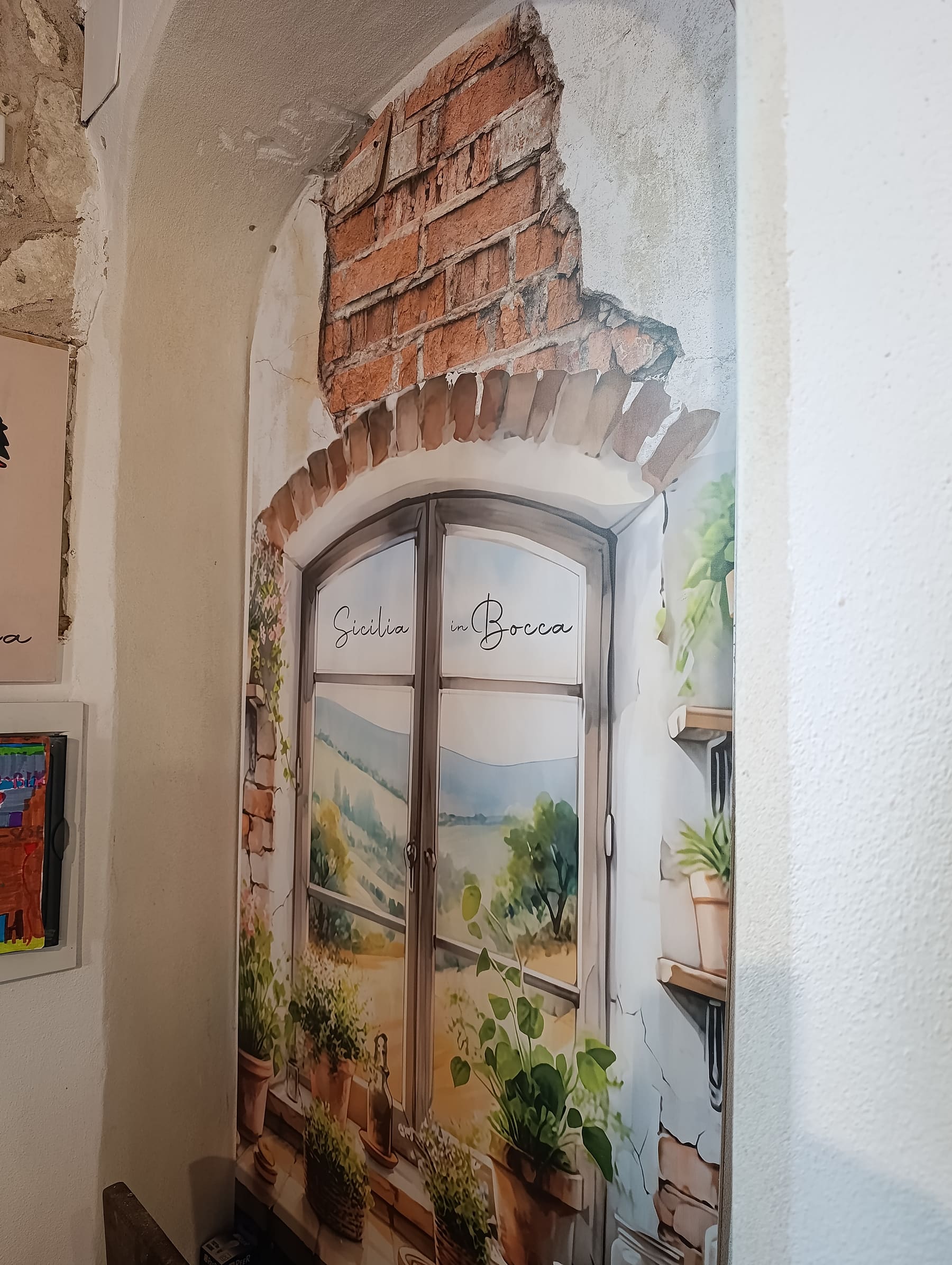 Murale trompe-l'œil con una finestra siciliana dipinta, mattoni a vista e vasi di piante, con la scritta a mano Sicilia in Bocca sui vetri