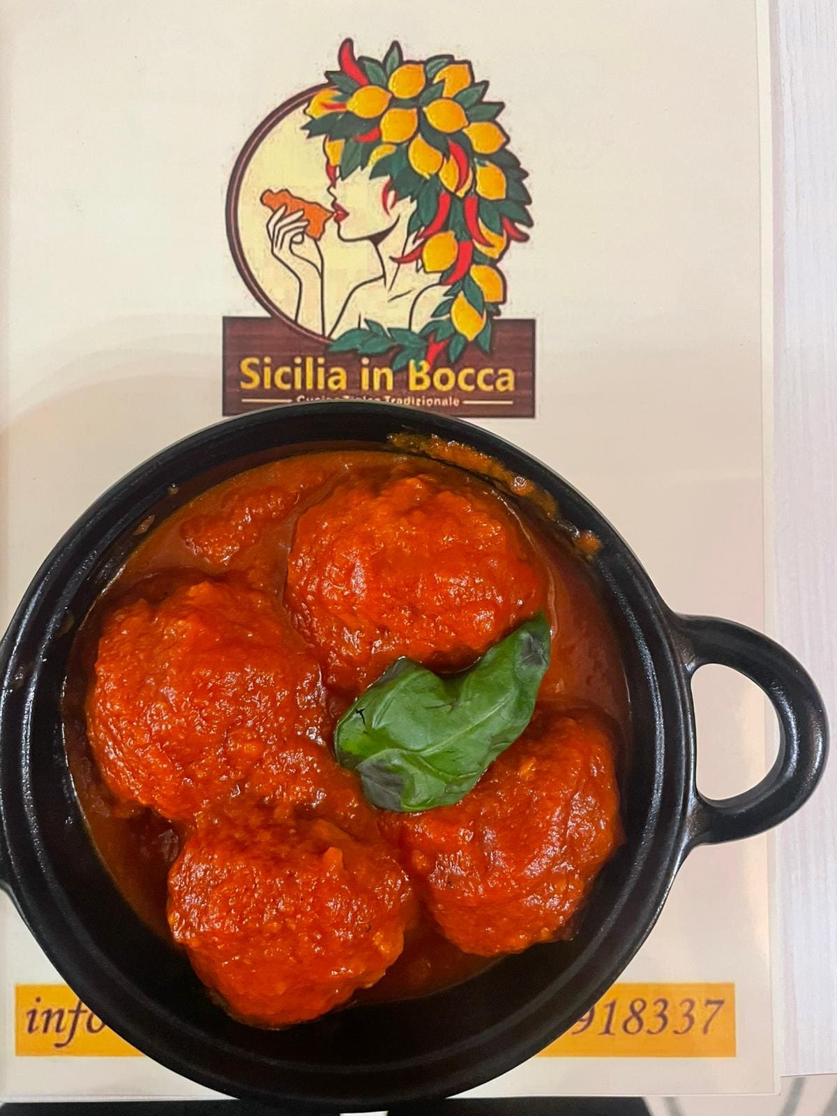 Polpette al sugo di pomodoro con una foglia di basilico, servite in un tegamino di ghisa appoggiato sul menu con il logo di Sicilia in Bocca